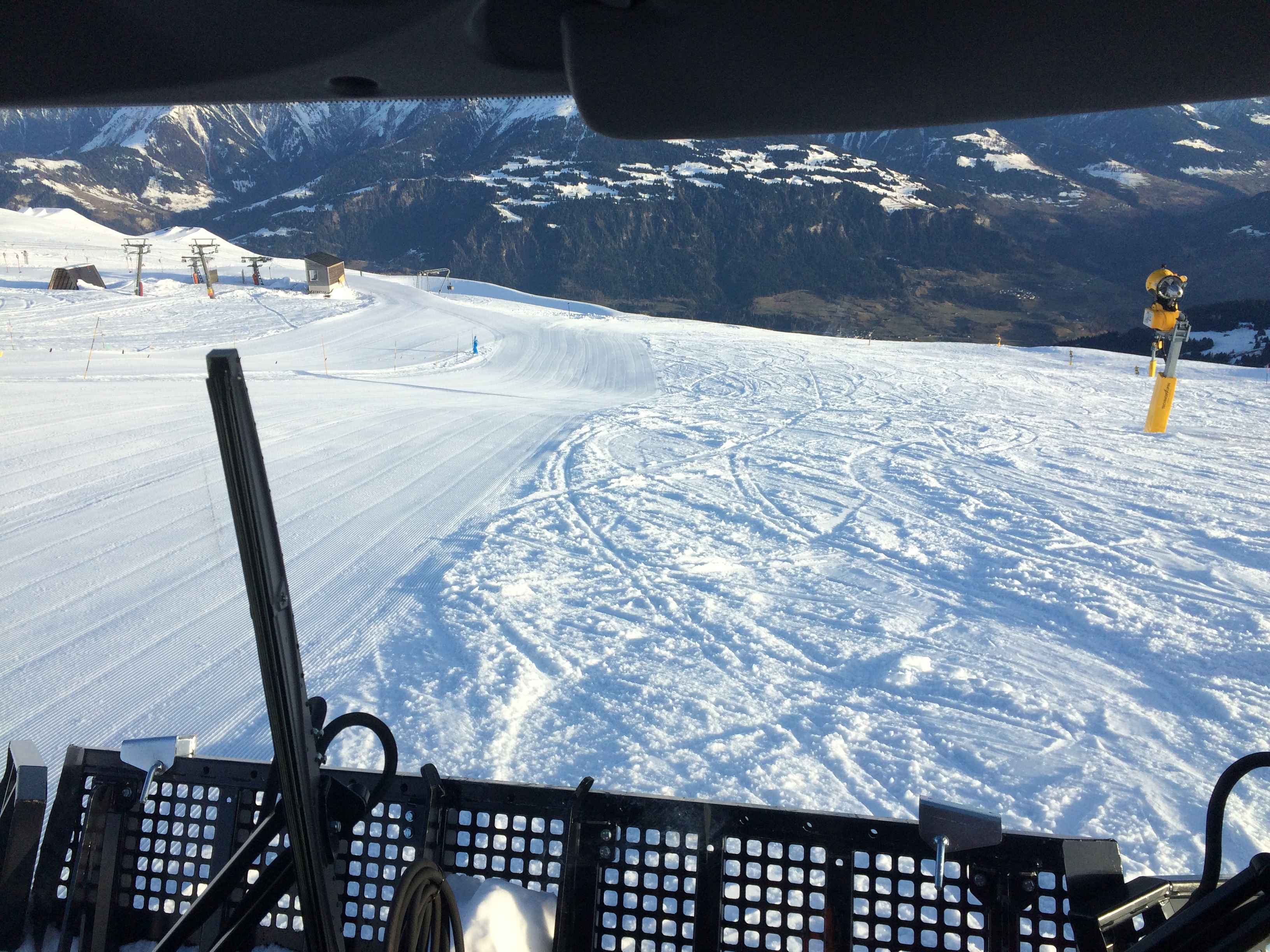 Blick aus der Fahrerkabine einer Pistenraupe über die Piste auf das Bergpanorama