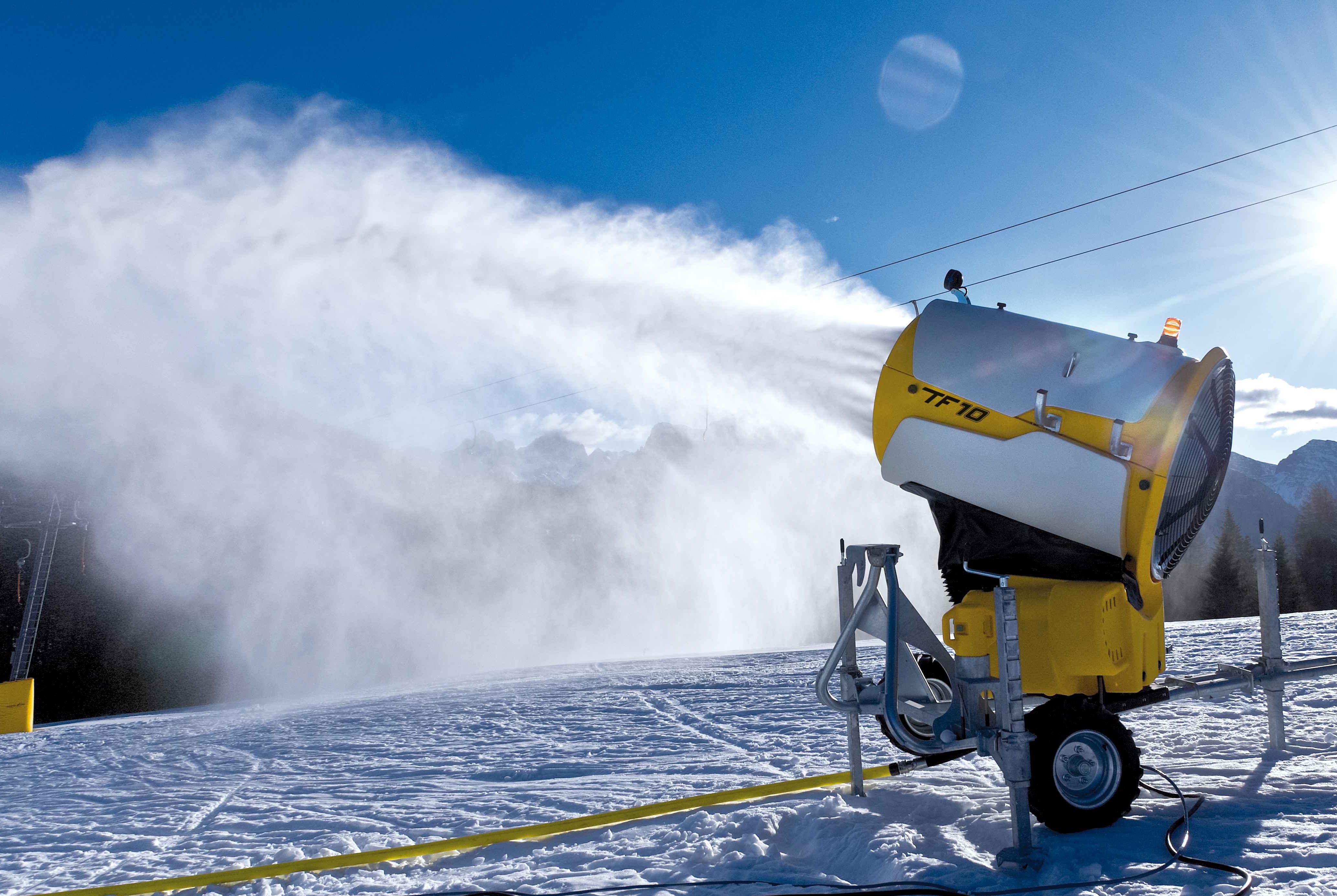 Schneekanone erzeugt technischen Schnee über einer Piste bei blauem Himmel